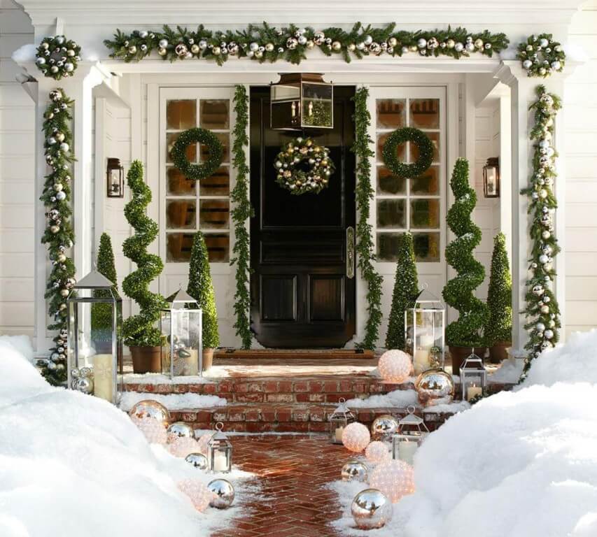 Front door to a home, covered with garland, wreaths, and Christmas decor all around, while a brick path to the door is just barely perceptible among the heavy snow