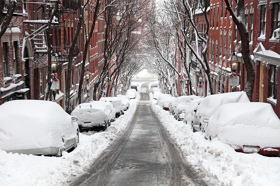 The city of Boston, Massachusetts in the winter. A scene of cars blanketed in snow, lining a narrow street that's been recently plowed.