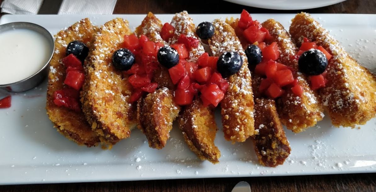crispy french toast with berries like blueberries and strawberries sprinkled with powdered sugar and frosting dipping sauce in a side container on a long white rectangular plate