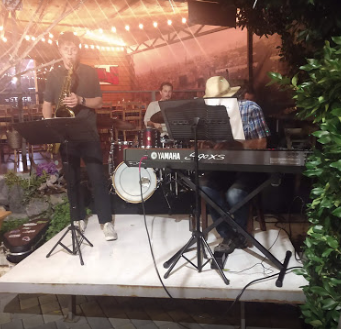 the stage cafe band playing on platform outside with dining and chairs and tables drum set saxophone piano