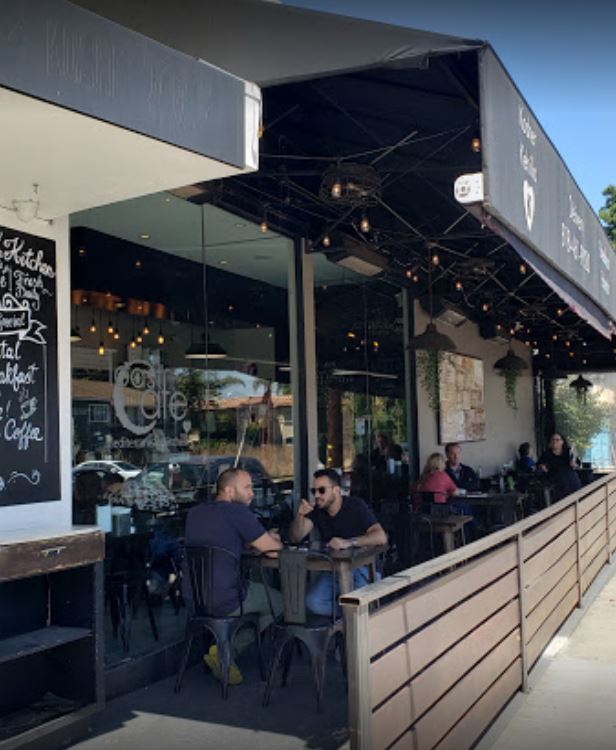 toast cafe outdoor dining area with wood looking gated eating metal chairs with wood tables chalk board black overhang lit up with string lights large glass windows looking in and people sitting and eating and drinking 