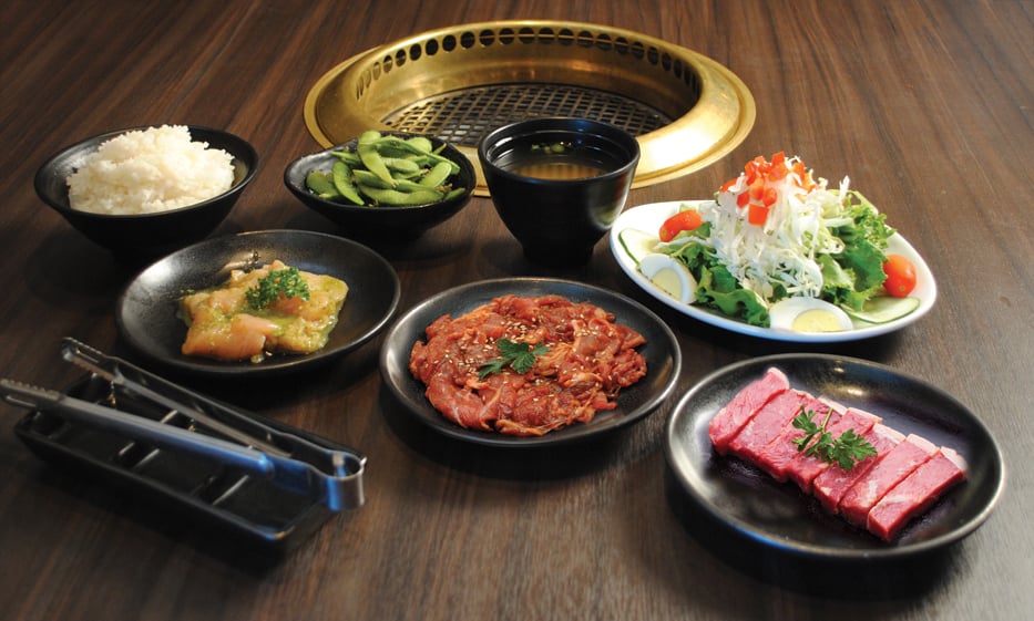 wood open grilling table with sauce plates tongs round black plates with raw meats bowl of rice white oval plate with salad edamame and a bowl of miso soup