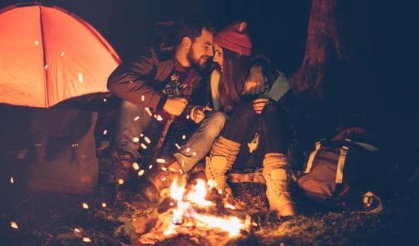 A happy, intimate couple presses their foreheads together as they are camping outdoors in front of a vibrant fire with an orange tent in the background. Both are wearing winter-weather clothes.