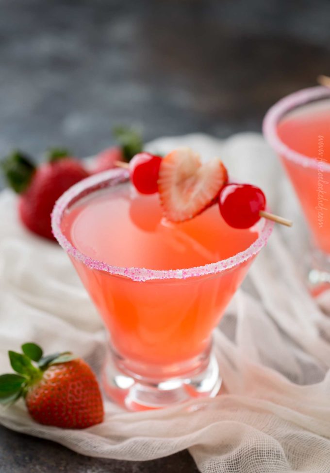 A vibrant orange-pink Valentine's Day cocktail known as love potion in a martini glass with a pink sugar rim garnished with maraschino cherries and one strawberry on a toothpick.