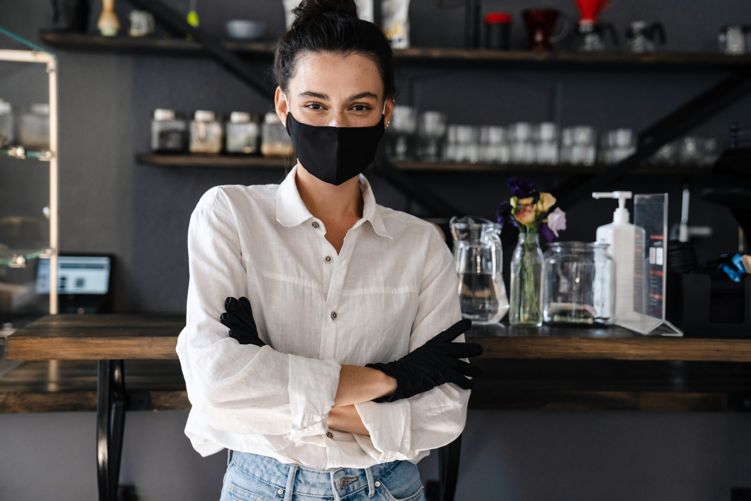 Female small business owner stands in front of her bar, wearing gloves and a face mask.