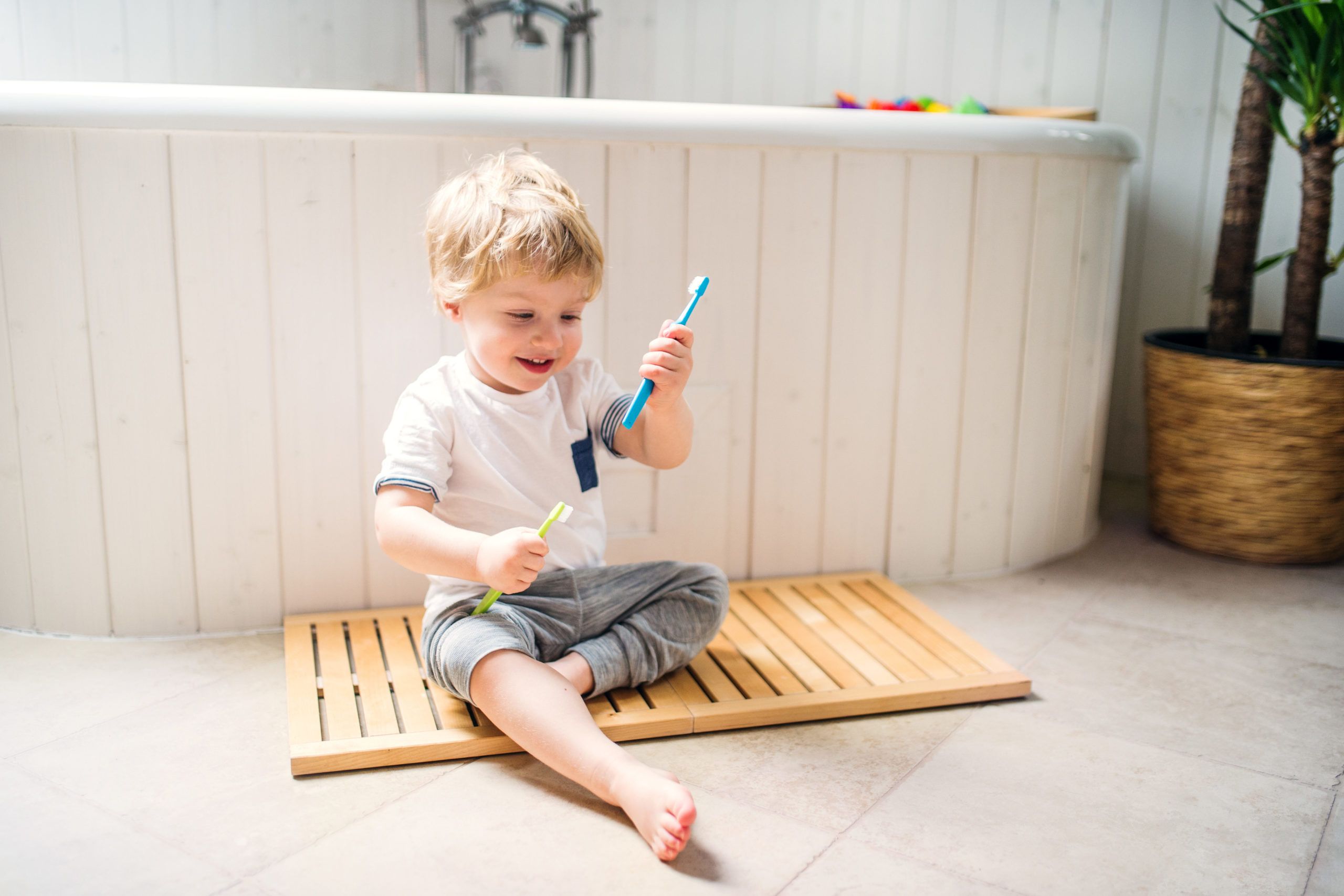 A happy toddler boy with a toothbrush sitting on the floor in the bathroom at home.