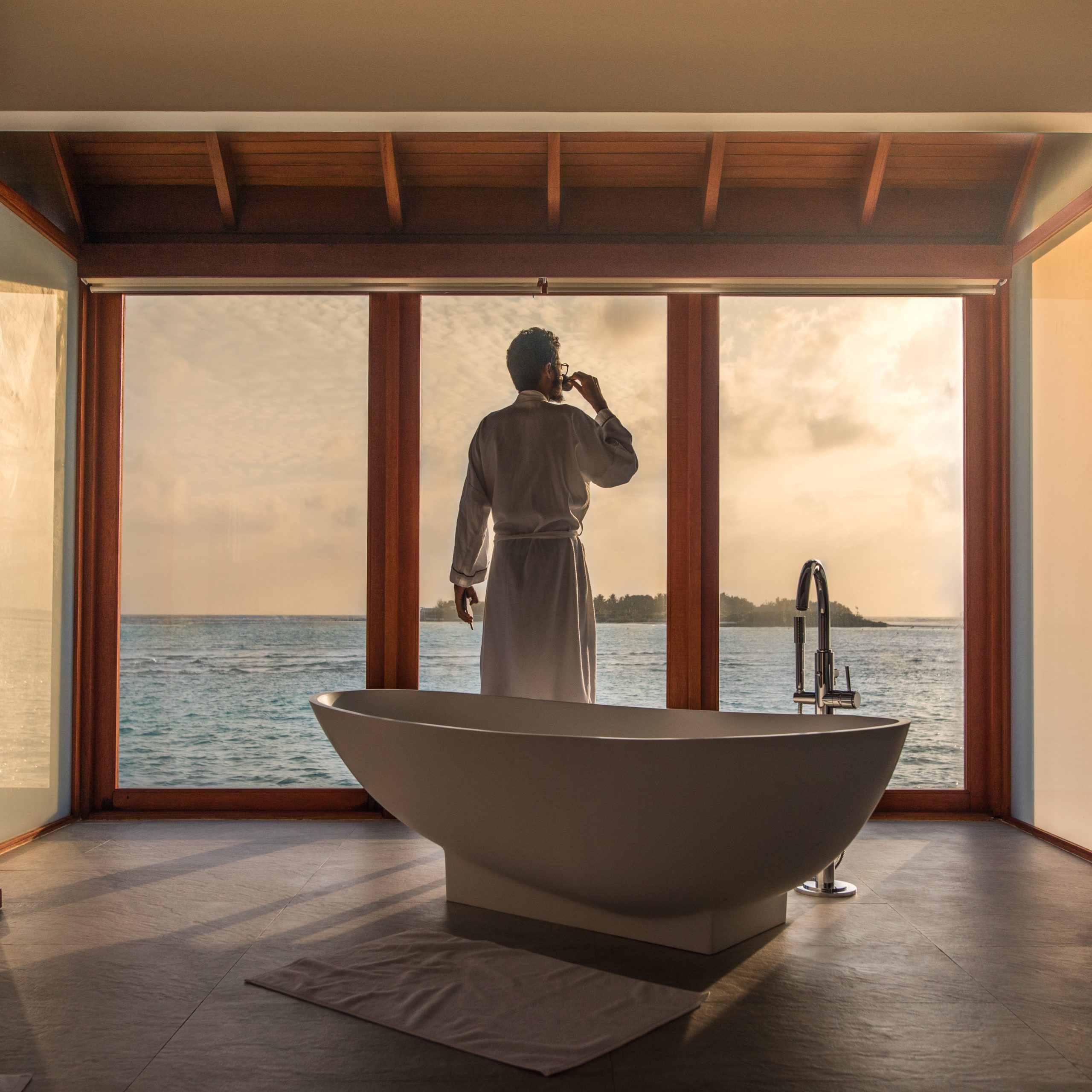 Man stands beside modern bathtub looking out the window at a body of water
