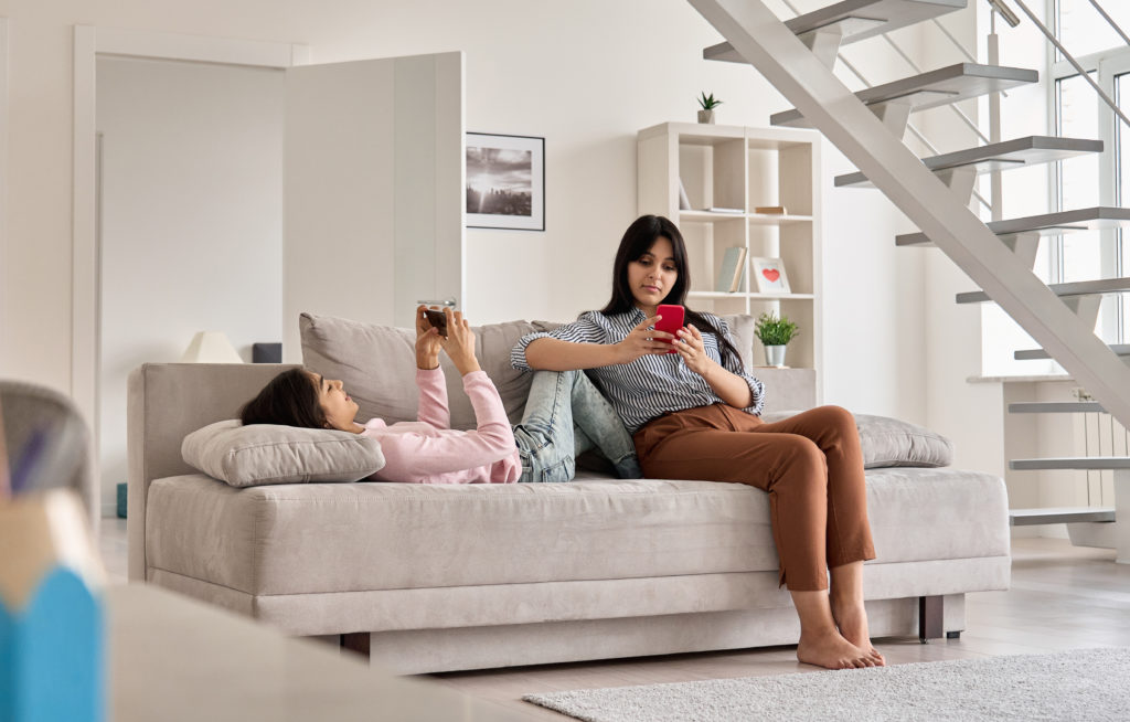 Indian family mother with teen daughter using phones devices at home. Young mom with teenage child holding cellphones relaxing together on couch in living room. Social media technology addiction.