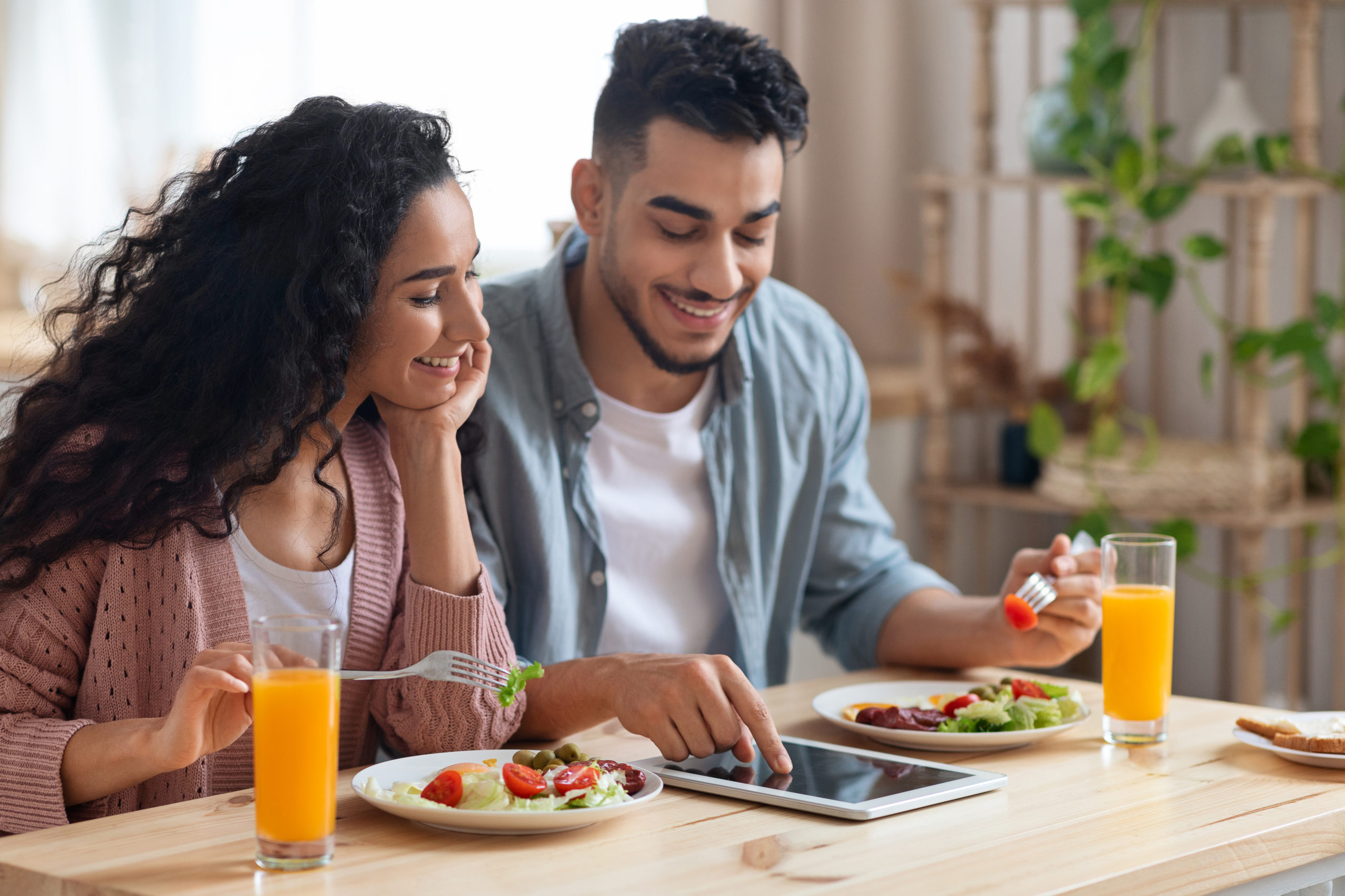 Joyful Middle-Eastern Couple Shopping Online With Digital Tablet While Having Breakfast In Kitchen, Young Arab Spouses Purchasing Grocery Delivery Or Browsing Internet While Enjoying Their Meal
