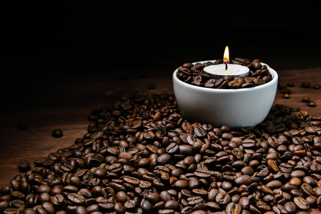 White coffee cup full of coffee beans and burning candle on top of coffee bean stack - image