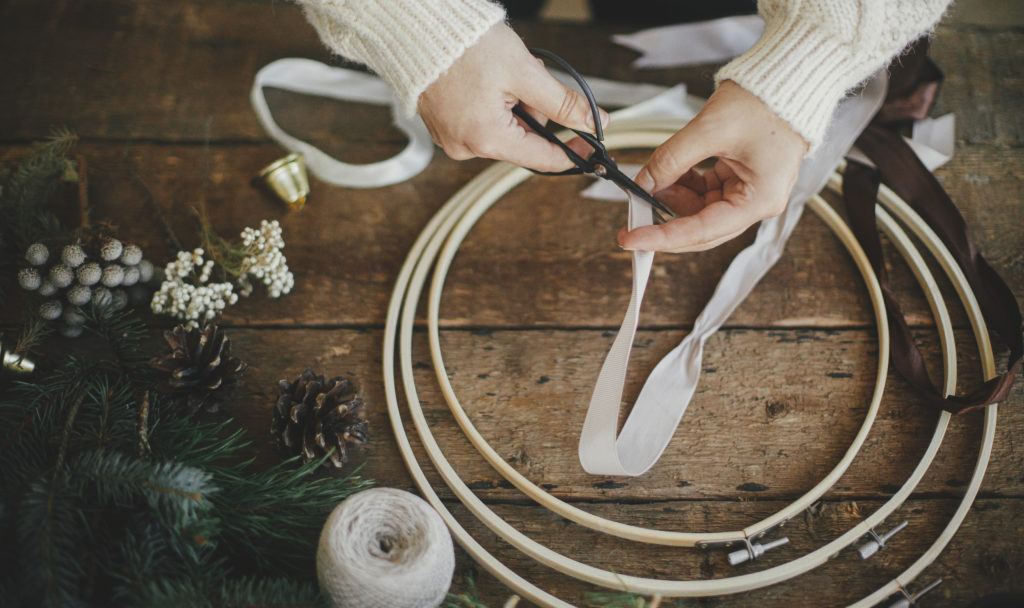 Making DIY decorations, stylish minimalist christmas wreath. Hands cutting ribbon with scissors for modern boho wreath with fir branches, brunia herb, wooden hoop on rustic table. Atmospheric moody image