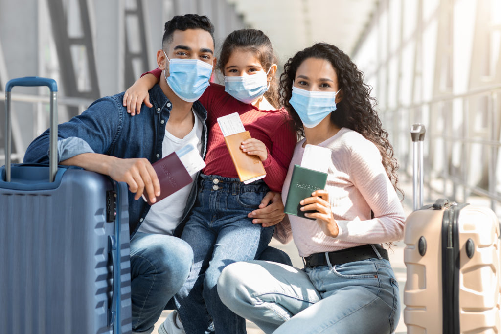 Family Travel During Pandemic. Parents And Little Daughter Wearing Medical Masks In Airport Waiting For Flight At Terminal, Sitting Near Suitcases With Passports And Tickets In Hands, Closeup