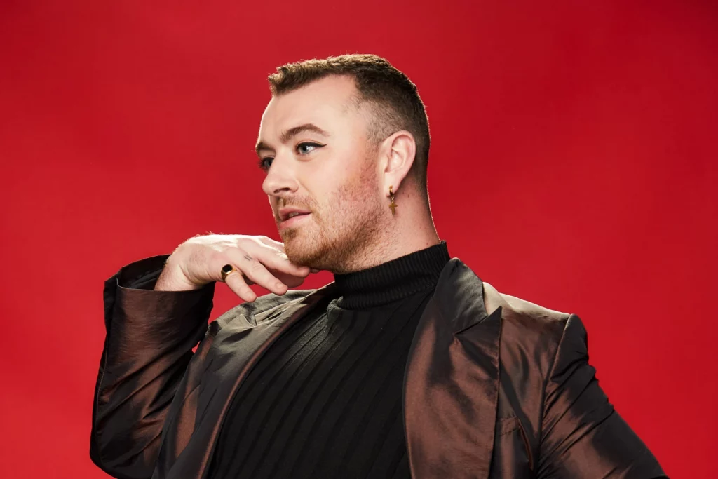 Sam Smith in brown jacket, black shirt, posing in front of a red backdrop
