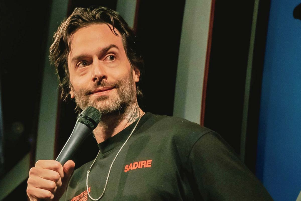 Comedian Chris D'Elia on stag holding a micophone to his mouth giving hard eyes to someone in the crowd. 