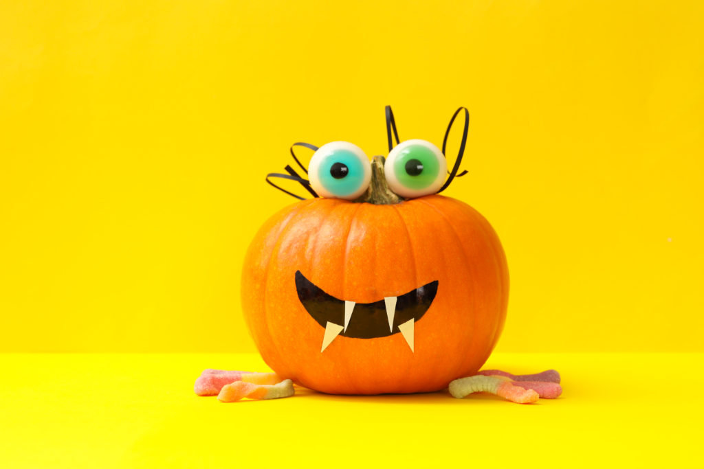 a decorated pumpkin with big green eyeballs placed on top of the pumpkin with sour gummy worms on a yellow background