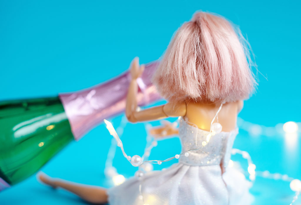 Barbie with light pink, short hair has her back to the camera as she faces a pink and green champagne bottle