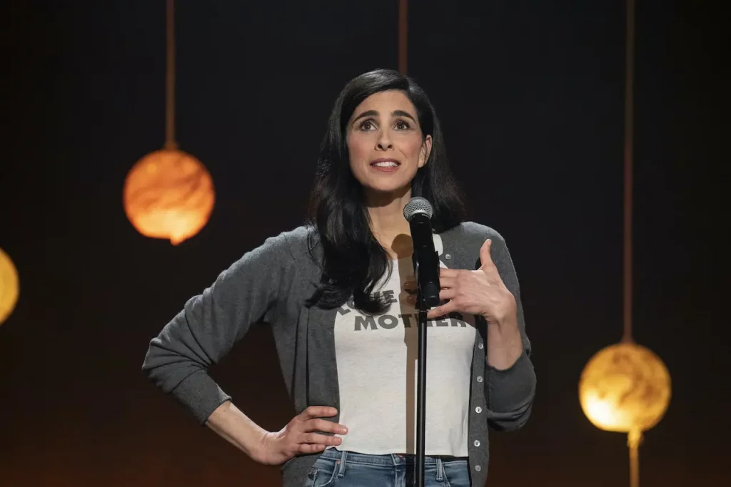 Comedian Sarah Silverman on stage wearing a white t-shirt and grey cardigan standing in front of a microphone with a hand on her hip. 