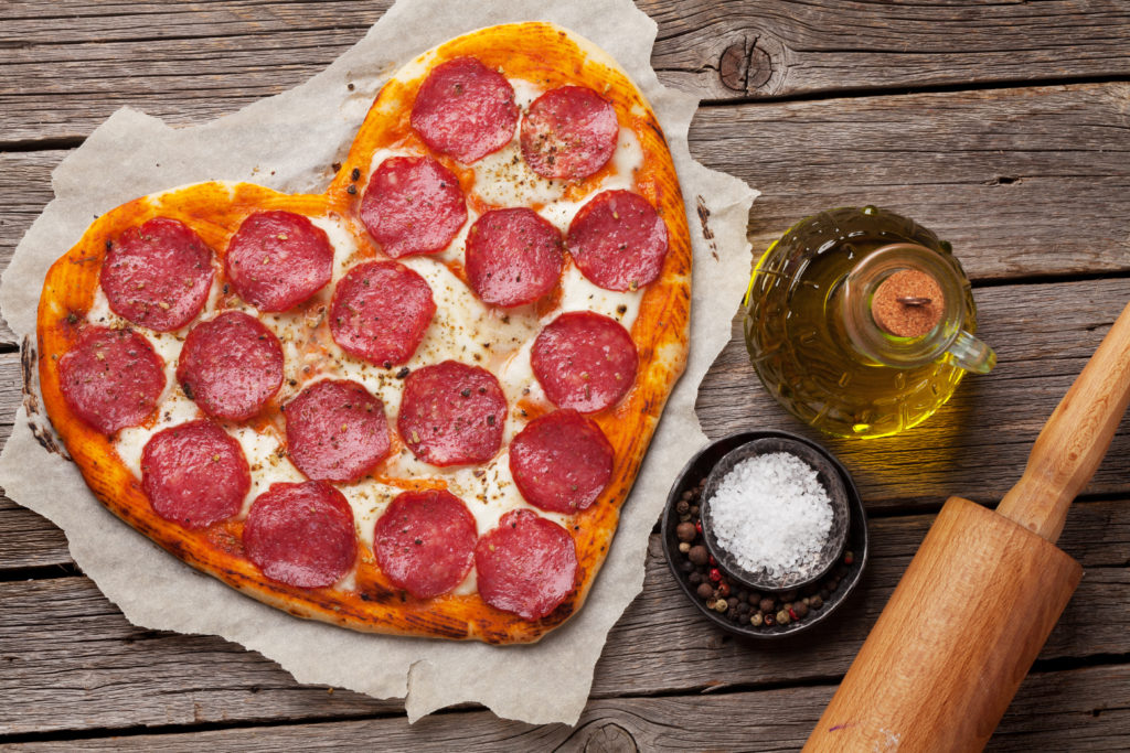 Fresh baked heart-shaped, Valentine pizza with pepperoni and various ingredients on wooden surface.