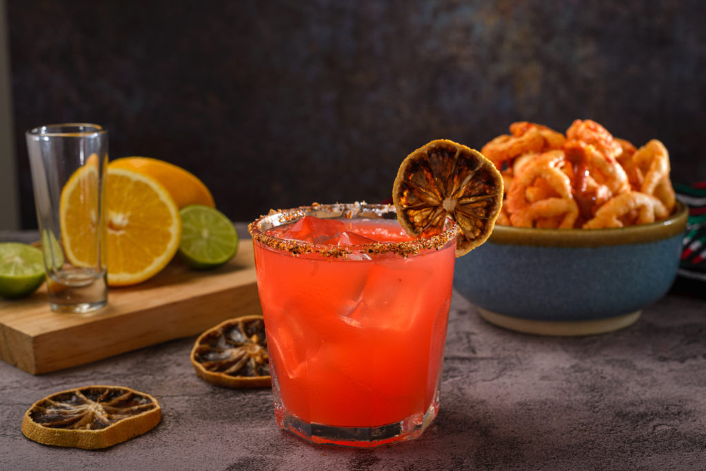 blood orange margarita with tajin rim and dried orange wedge with various items in the background.