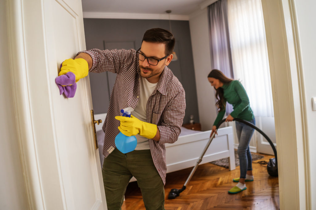 Young Caucasian couple cleaning a bedroom, vacuuming and wiping door with spray and towel.