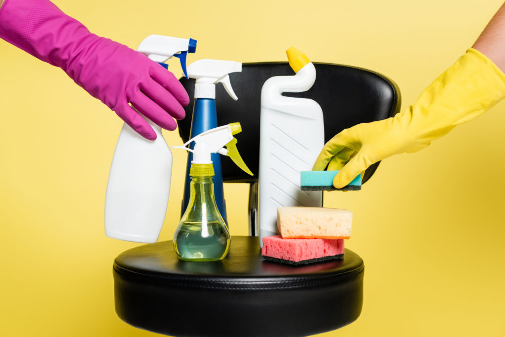 Two hands, one with pink glove, and another with yellow glove placing cleaning items on a chair along with other cleaning supplies on yellow background.