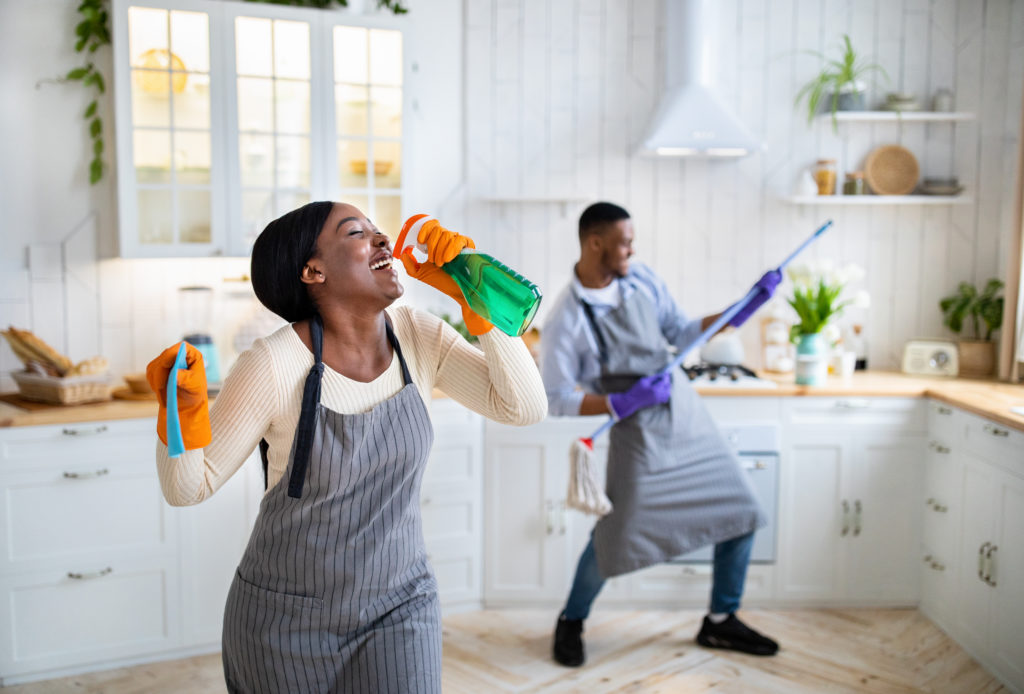 Young black couple in kitchen using cleaning supplies to act out singing and guitar playing.