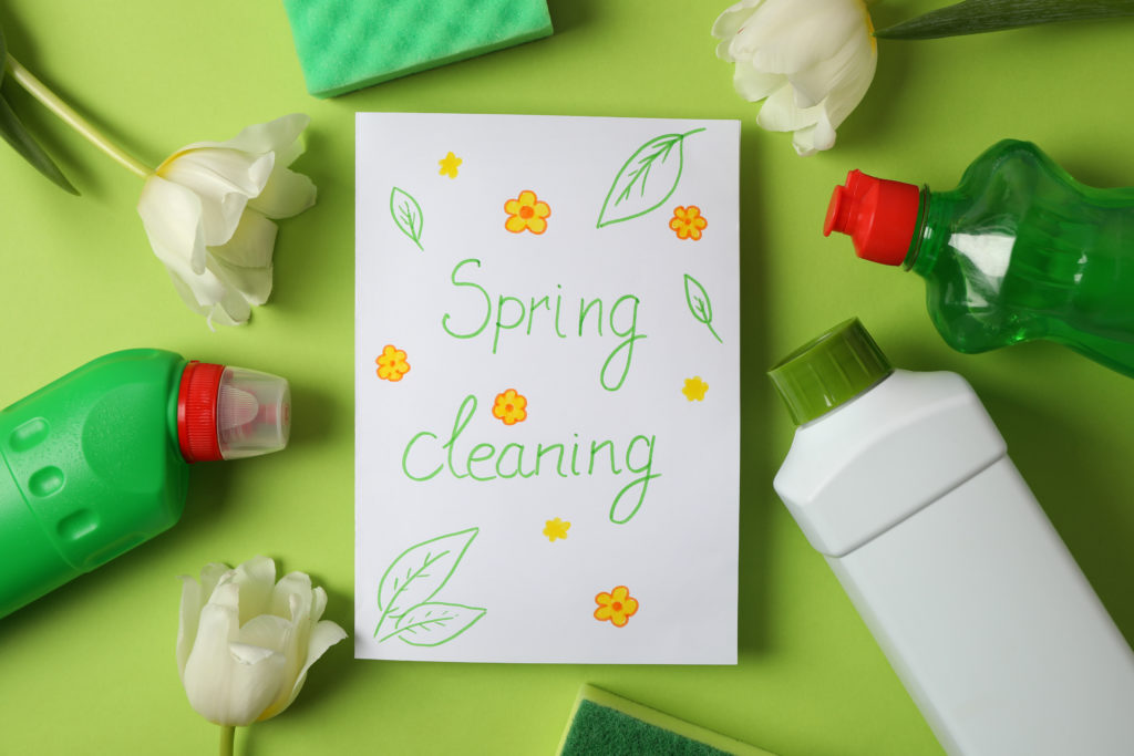 paper that reads "Spring cleaning" with flowers and leaves drawn and surrounded by cleaning products on a green background.