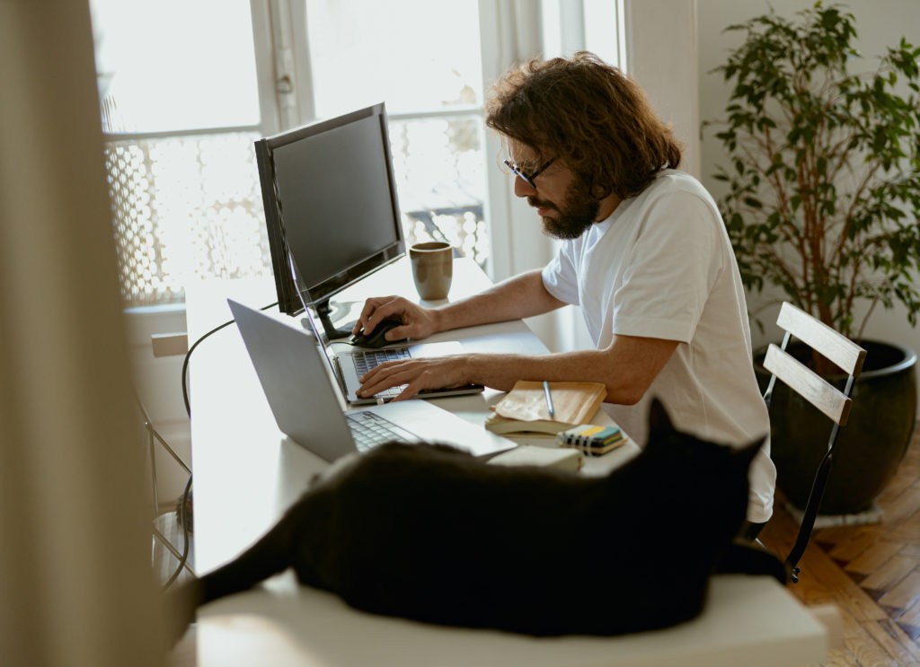 Bearded man with glasses working on two laptops and one desktop computer.